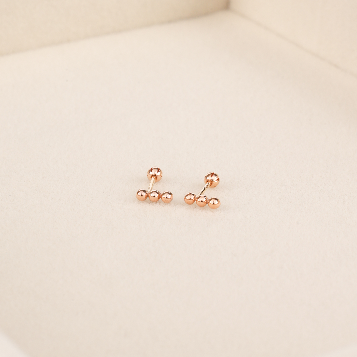Dainty rose gold bar stud earrings with three ball details and rounded screw-back posts on beige surface