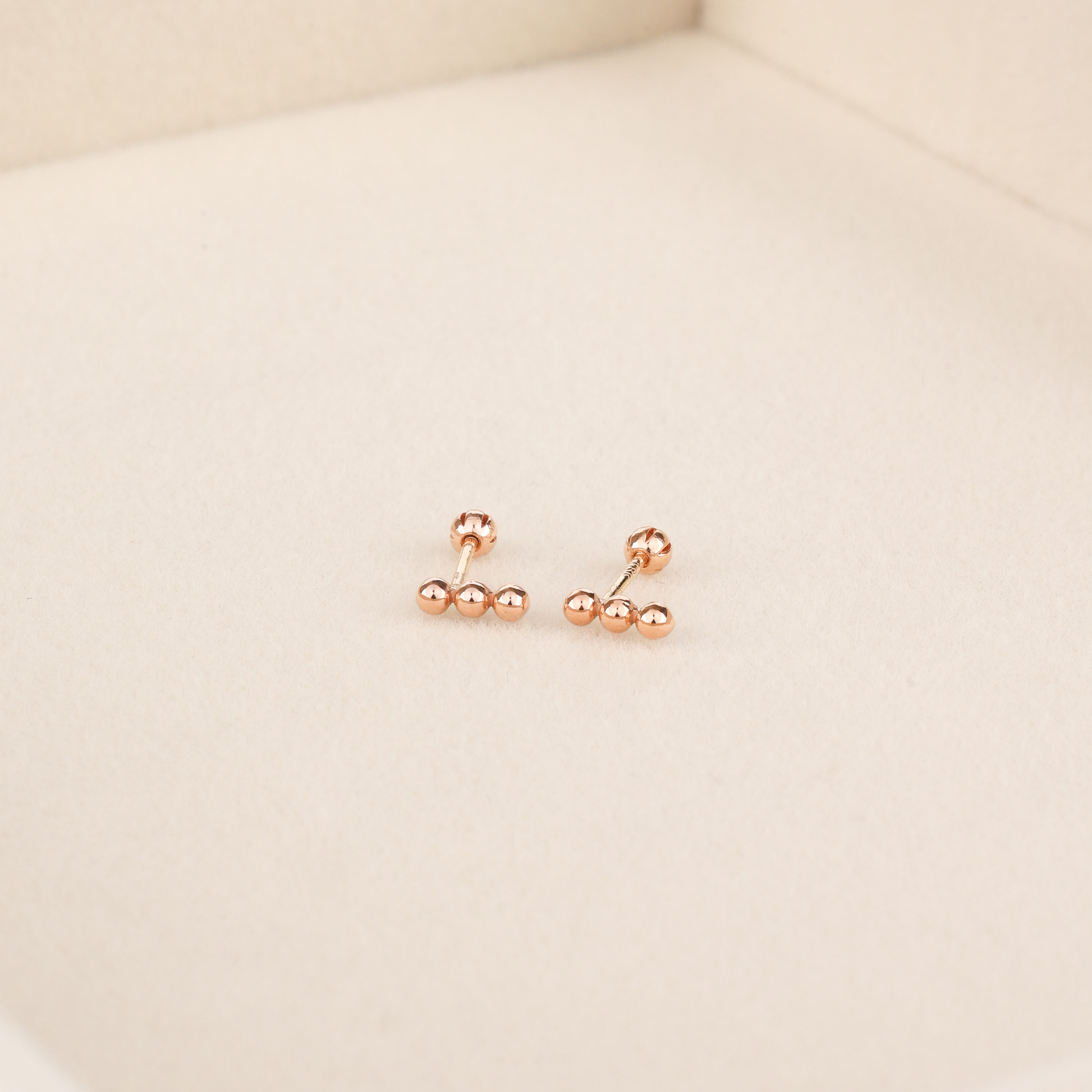 Dainty rose gold bar stud earrings with three ball details and rounded screw-back posts on beige surface