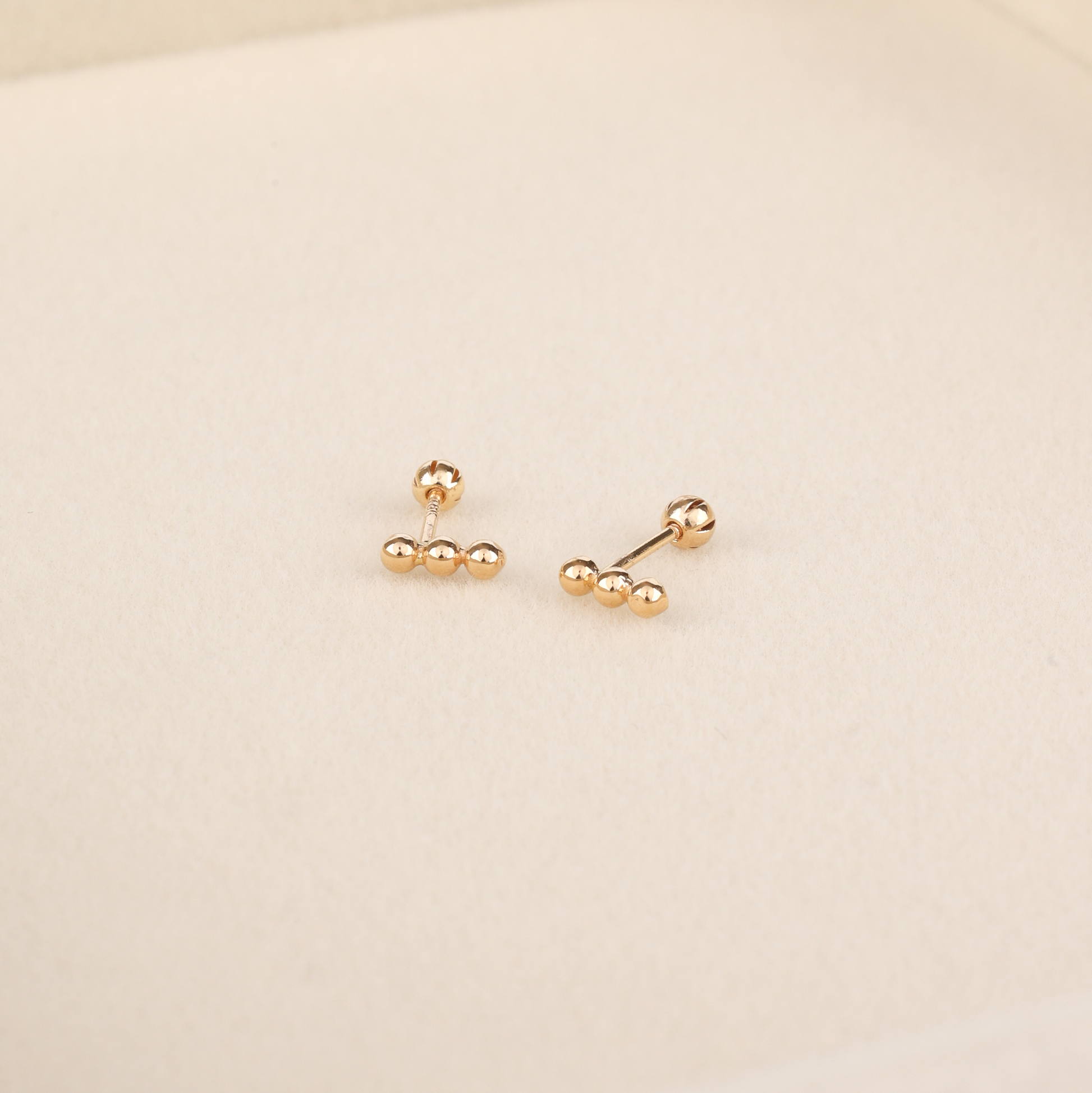 Dainty yellow gold bar stud earrings with three ball details and rounded screw-back posts on beige surface
